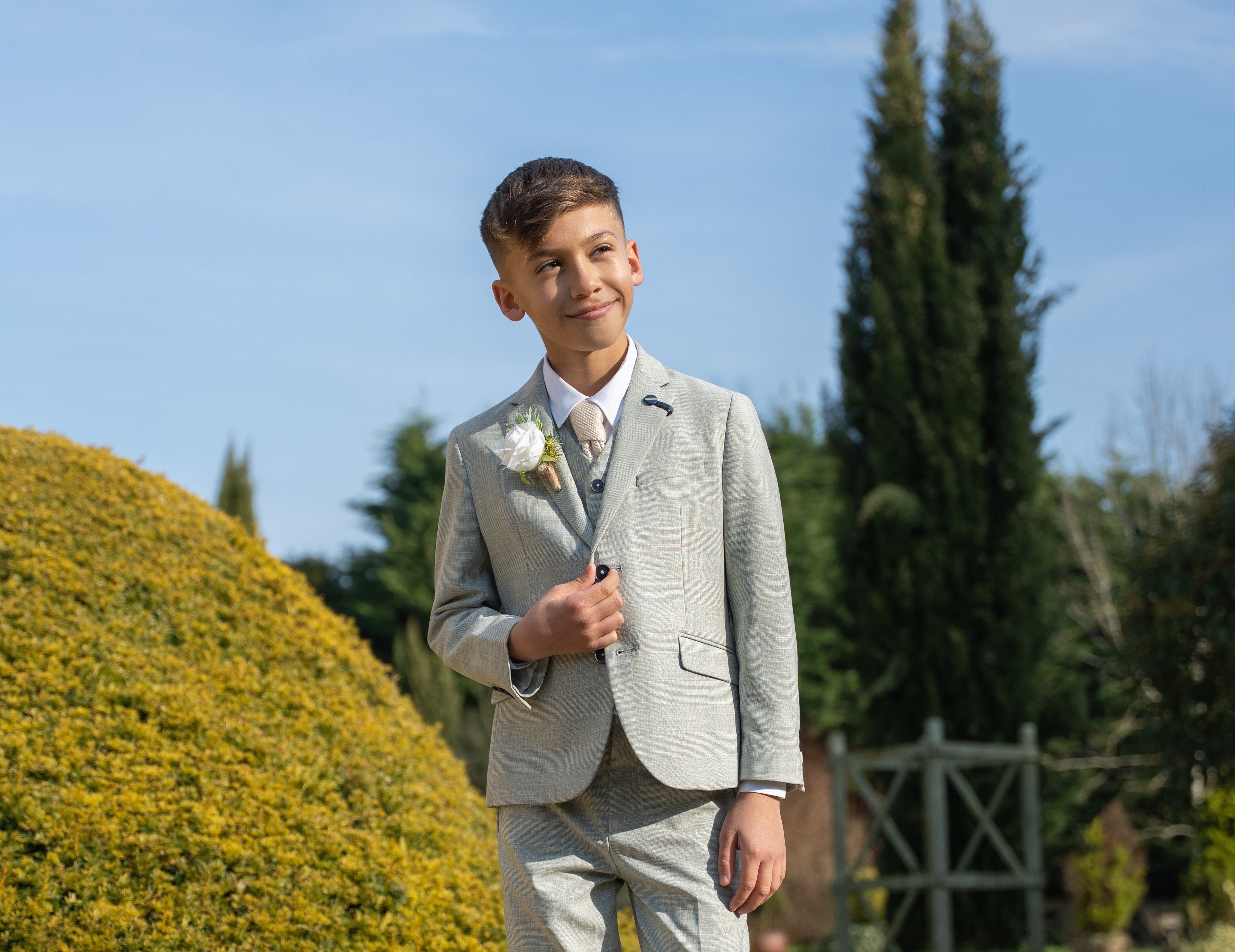 Boy in a green summer suit with boutonnière, standing outdoors on a sunny day, smiling and looking to the side.
