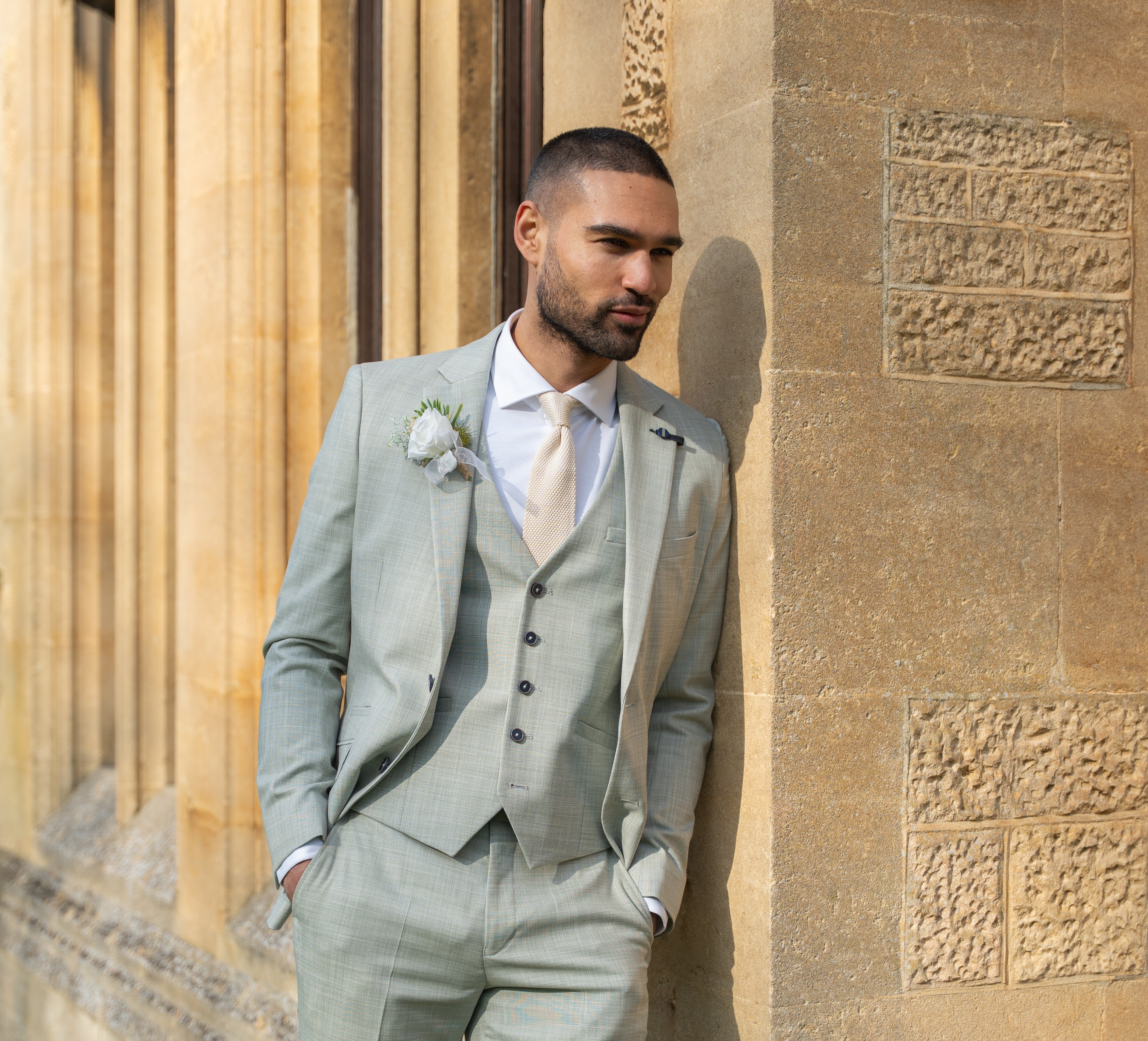 A man standing against a wall wearing a sage green summer wedding suit