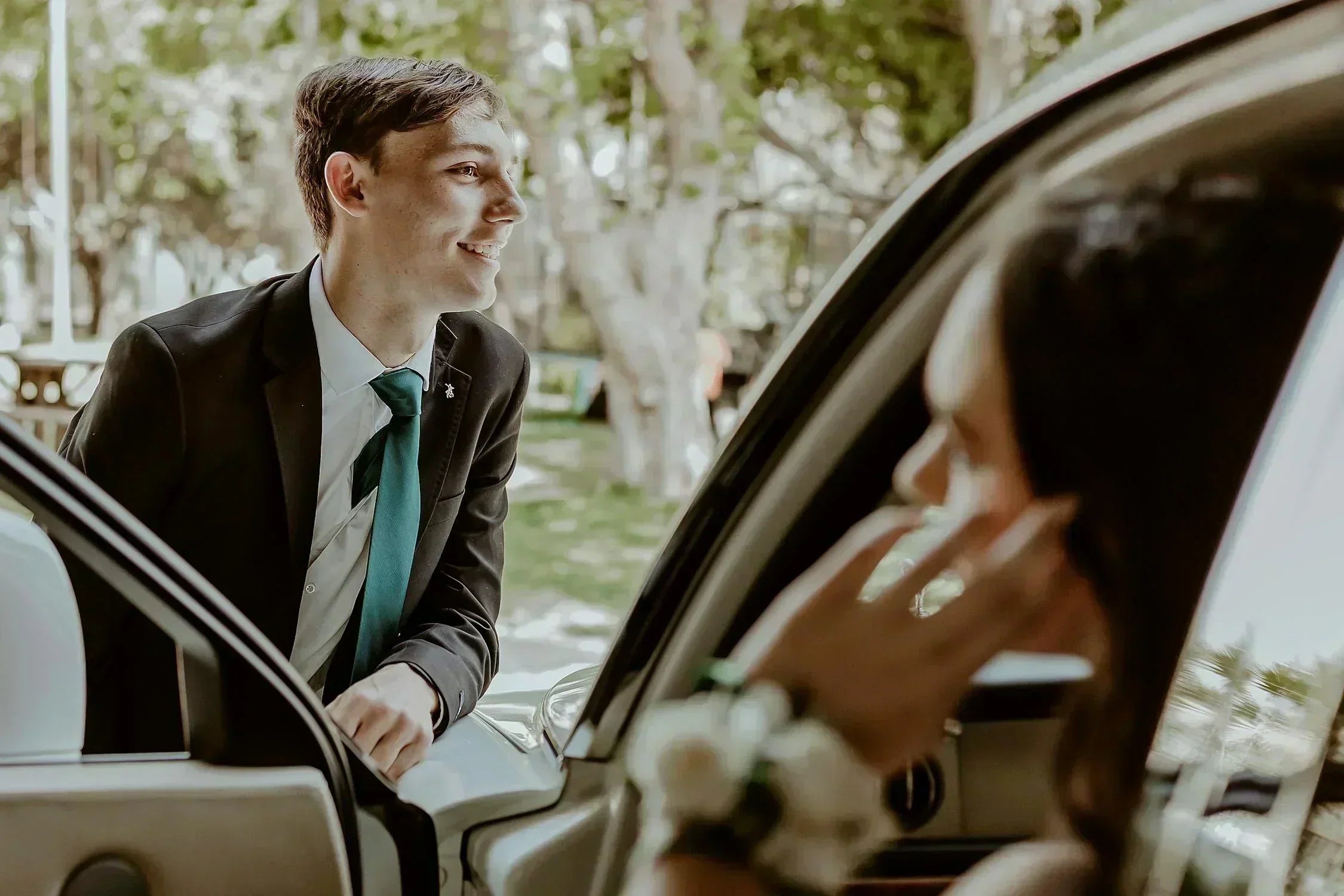 a young man in a prom suit standing by the door of a car and young woman sitting inside of the car